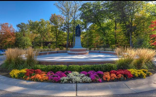 圣雕像. Thomas Aquinas at the center of a paved circle surrounded by a short brick wall lined with bushes, 花, 和草. 背景是一排排的树.