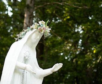 法蒂玛圣母 Statue adorned with beads and a flower crown with a backdrop of green tree foliage.