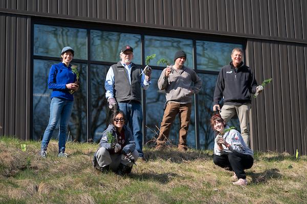 Six AQ community members (staff, faculty, students), smile and hold tree saplings on a hill near Sturrus