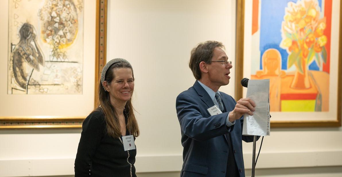 Aurelia Engel, daughter of Francoise Gilot stands beside Jochen Wierech who speaks into a microphone in the gallery with Gilot works behind him.